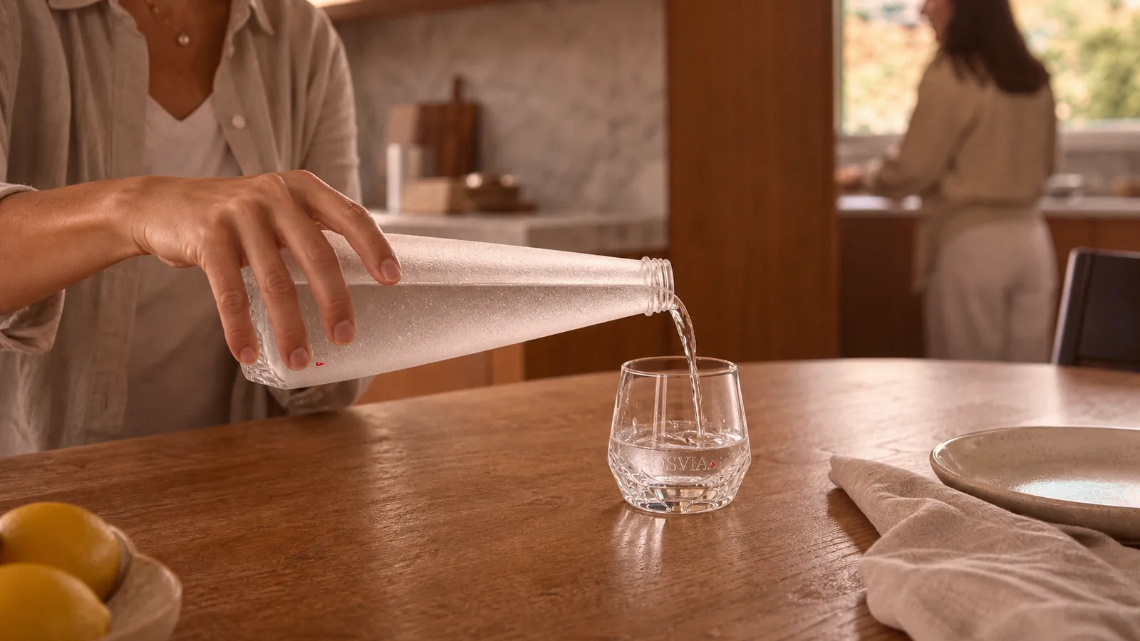 Une main sert de l'eau depuis la bouteille OSVIA givrée dans un verre gravé, sur une table de cuisine en noyer baignée de lumière du jour, un membre du foyer en arrière-plan